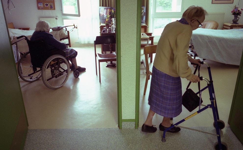 Til venstre sidder en ældre dame i kørestol. En væg skiller hende fra en anden dame, der er på vej ind på et værelse med sin rollator.