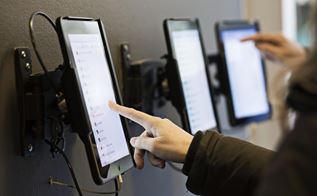Tre iPad's hænger på en væg, mens to personer står og trykket på to forskellige skærme med deres pegefinger.