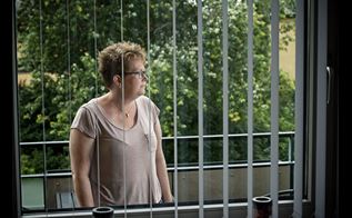 Tidligere Sosu-hjælper Jeannette Stripp stående på sin terrasse, fotograferet gennem vinduet