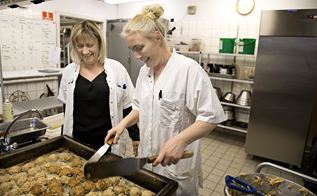 Britta Larsen står i et industrikøkken og steger frikadeller, iført hvid uniform med navneskilt