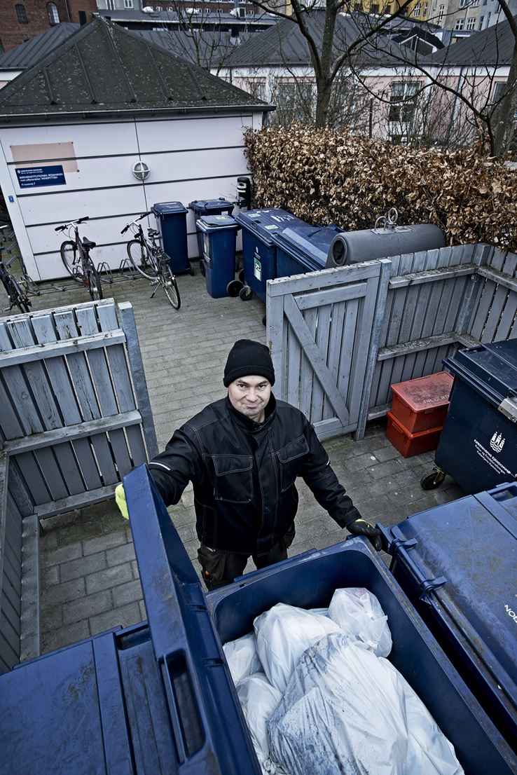 En mand holder i låget på en affaldscontainer, og smiler op mod kameraet