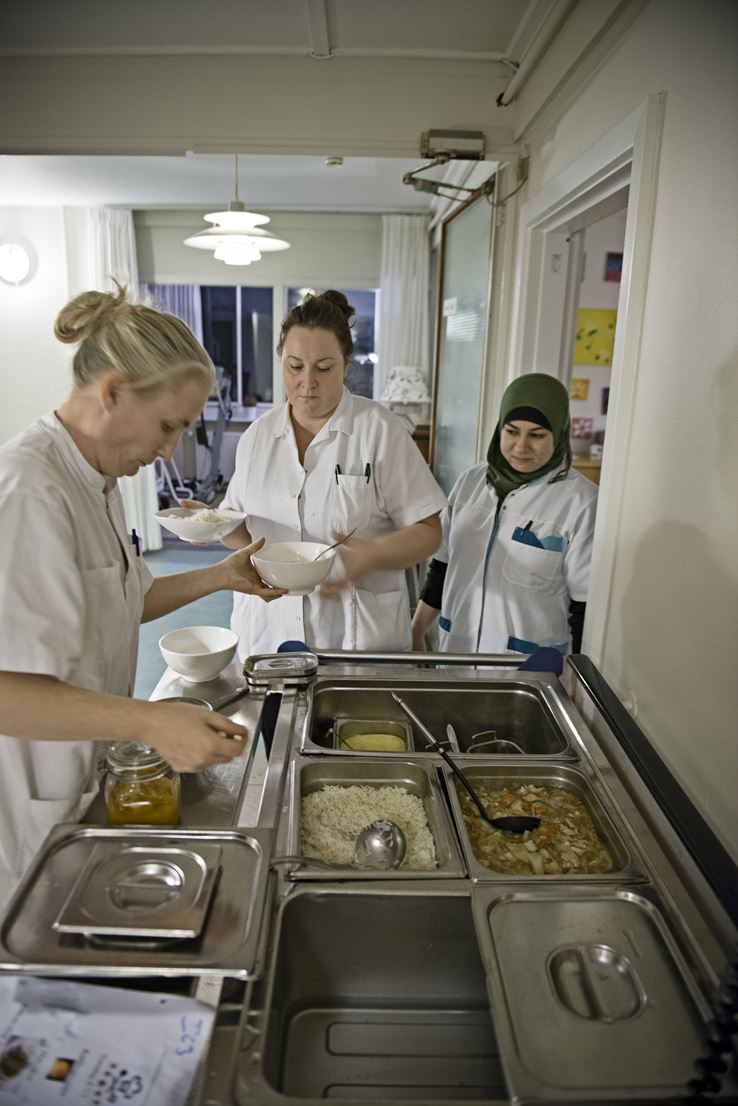 Britta Larsen står ved siden af madvognen, hvor hun øser op med den ene hånd og giver skålen videre til de ansatte med den anden. Fatme står klar til at modtage en skål. 