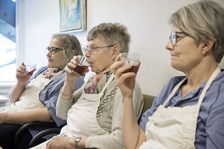 Tre kvinder sidder på række og drikker, iført hvidt forklæde og briller