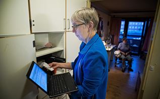Kvinde i blå trøje og med briller står og kigger på en computerskærm ved indgangen til en ældrebolig.
