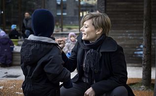 Trine Hermansen sidder på hug ved siden af et barn, med en legeplads i baggrunden