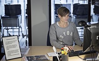 Trine sidder ved et skrivebord foran sin computer, iført en grå trøje med skriften "customer", ved siden af hende står et indrammet dokument med overskriften "Charlottehavens DNA"