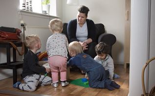 En kvinde sidder i en stol, og kigger ned på fire børn som ligger og leger på gulvet foran hende