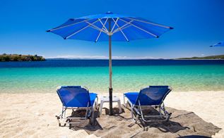 Strand med to blå strandstole og en blå parasol som vender mod blå-grønt hav.