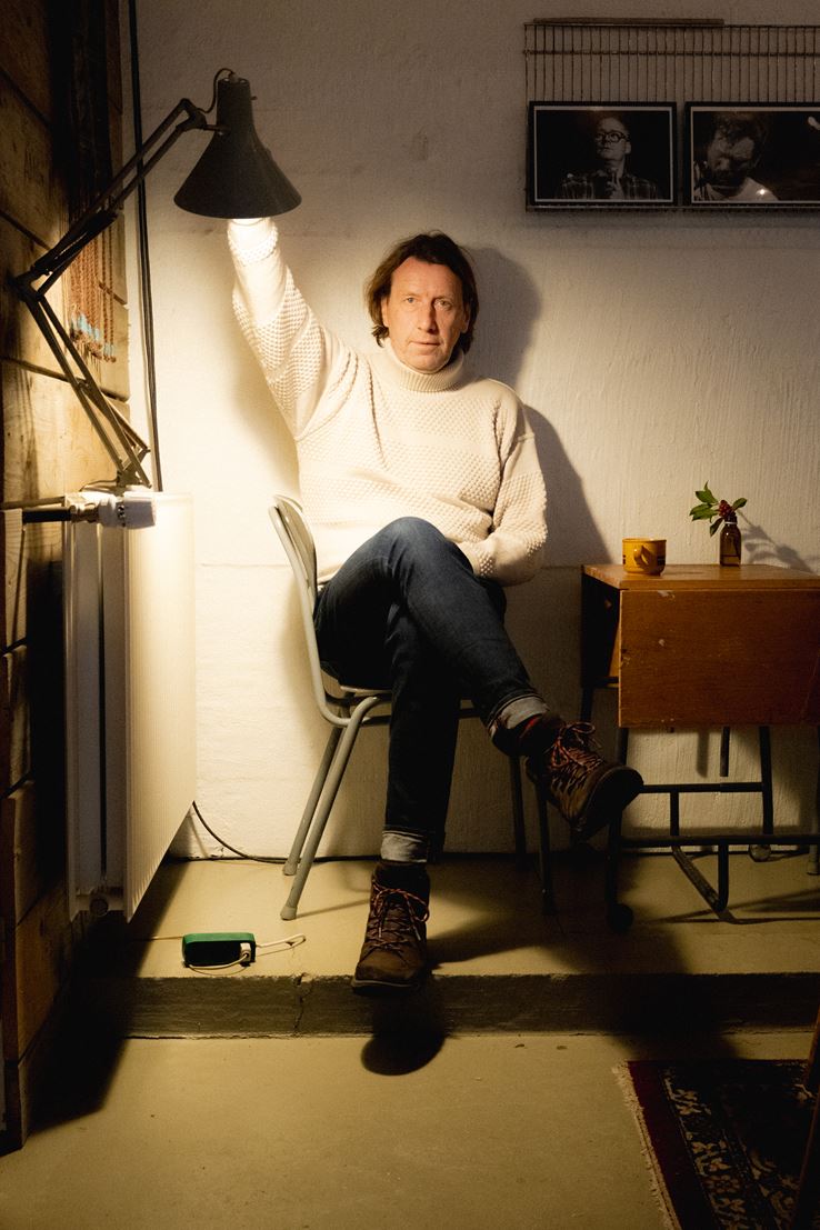 Anders Agger sidder på en stol ved et lille bord. Han holder ved en arkitektlampe, som lyser ham op.
